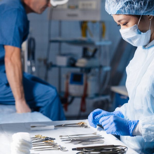 Asian female nurse looking at surgical steel medical instrument for group of surgeons at background operating patient in surgical theatre.